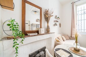 Una habitación con chimenea, espejo, planta, florero con plantas secas, una mesita con un vaso y ventana con cortinas.