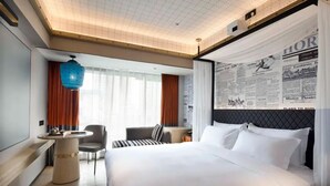 Ein Hotelzimmer mit einem großen Bett, einem runden Tisch, einem Stuhl, einem Schreibtisch und einem großen Fenster mit Vorhängen.