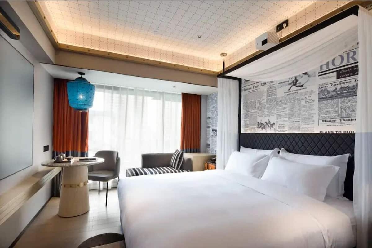 Habitación de hotel con una cama grande, una mesa redonda, una silla, un escritorio y un ventanal con cortinas.