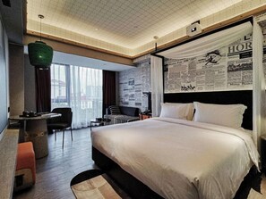 Une chambre d’hôtel moderne, dotée d’un grand lit, d’un petit bureau, d’une chaise et offrant une vue sur la ville grâce à de grandes fenêtres.