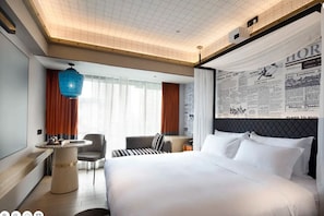 A hotel room with a large bed, a desk, a chair, a round table, and a blue hanging light fixture.