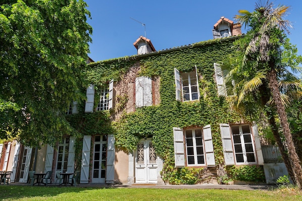 Un bâtiment aux murs recouverts de lierre, doté de grandes fenêtres avec volets et d’une cheminée bien visible.