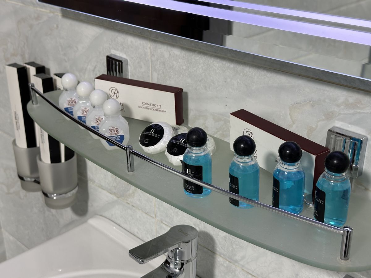 Un bany amb una prestatgeria de vidre que conté diversos productes de tocador, incloent ampolles de xampú i condicionador, i una caixa amb la inscripció "COSMETIC KIT".