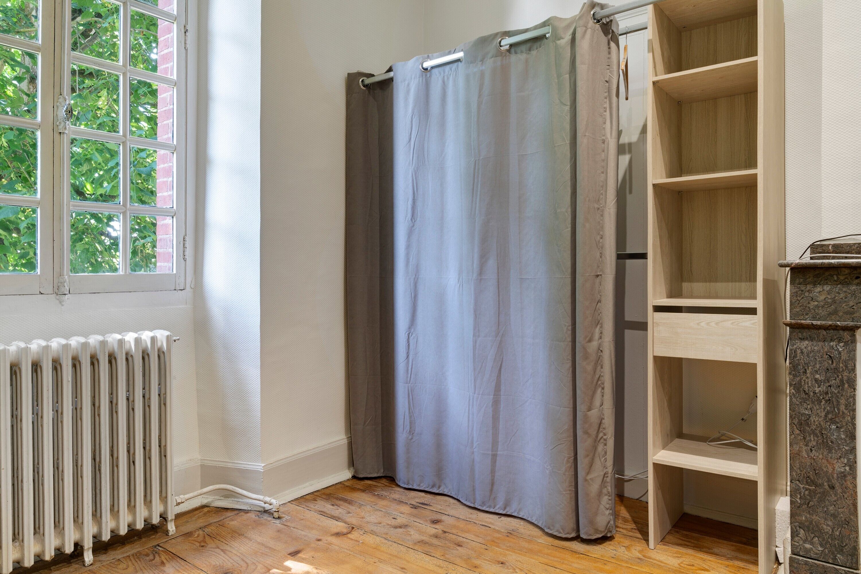 A room with a radiator, a window with a view of trees, a grey curtain, and a wooden shelf unit.
