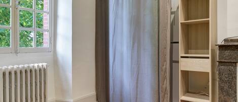 A room with a radiator, a window with a view of trees, a grey curtain, and a wooden shelf unit.