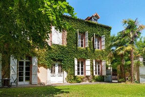A two-story building with a brick facade partially covered in green ivy, white shutters, and a small balcony.
