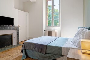 Une chambre Ă coucher avec un lit, un tĂ©lĂ©viseur posĂ© sur un meuble, une fenĂȘtre donnant sur des arbres et un radiateur.