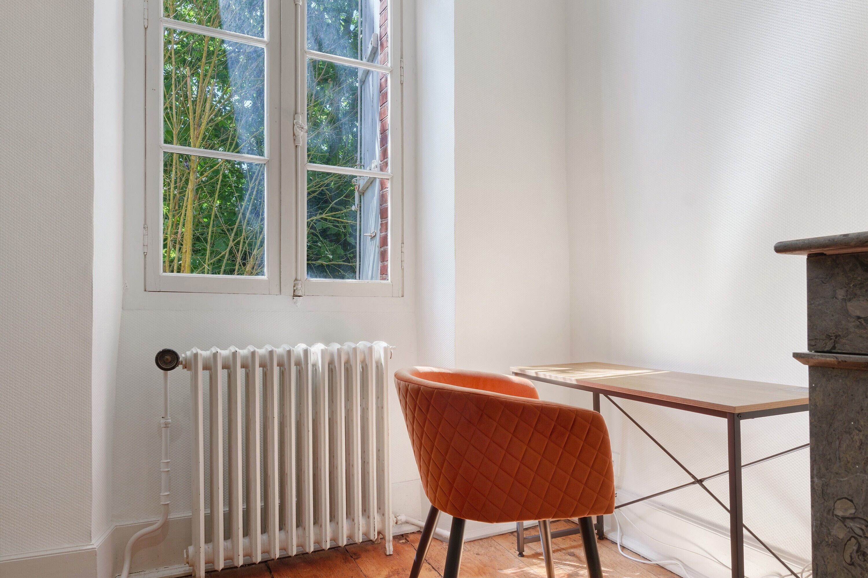 Ein Zimmer mit Heizkörper, einem Fenster mit Blick auf Bäume, einem Tisch und einem orangefarbenen Sessel.