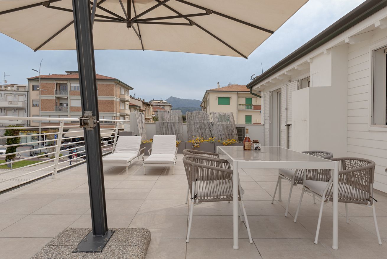 Une terrasse extérieure avec une table et des chaises, un grand parasol, et une vue sur les bâtiments et les montagnes en arrière-plan.