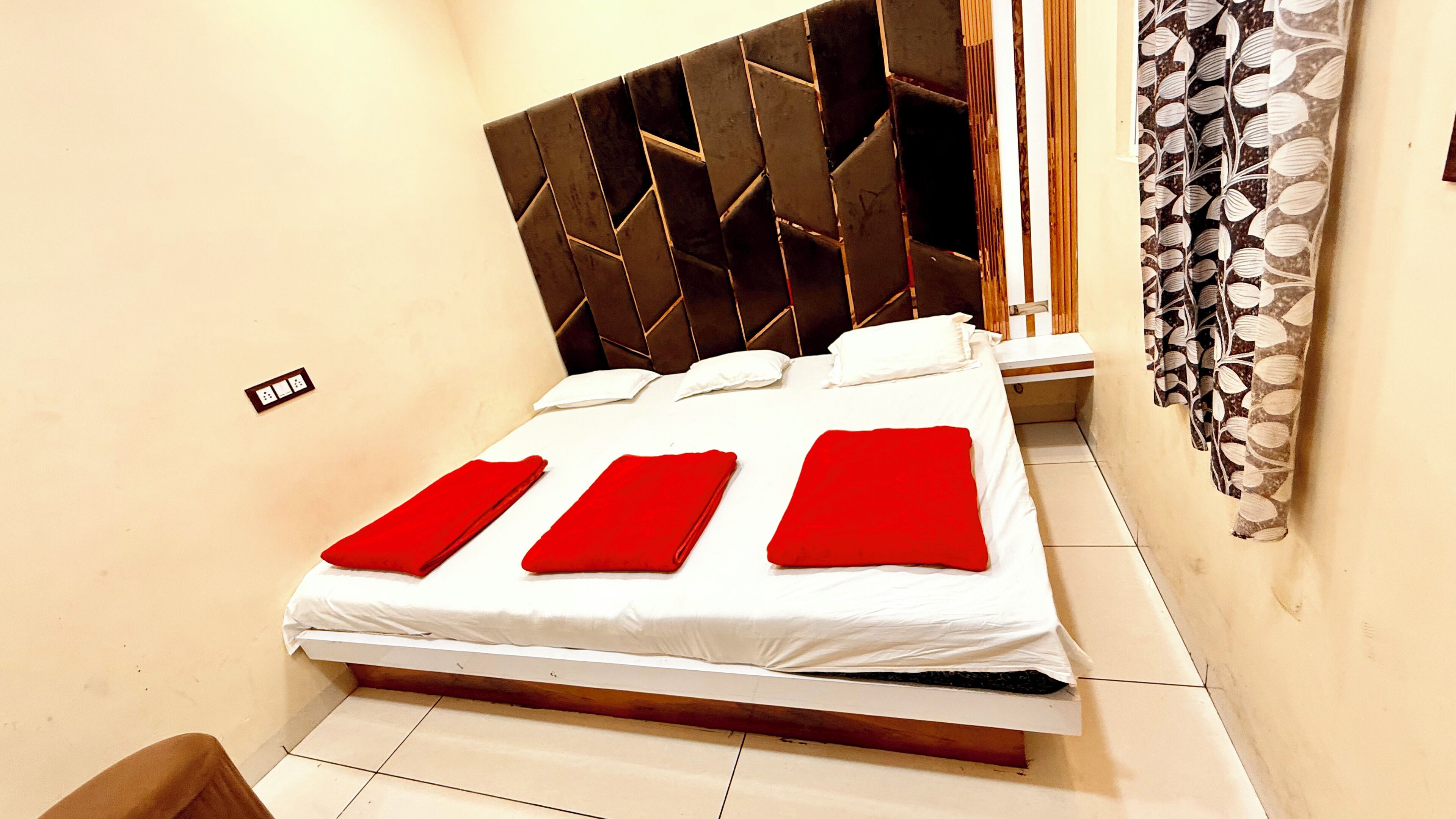Una cama bien tendida con sábanas blancas y almohadas rojas, un cabecero moderno y una mesita de noche pequeña.