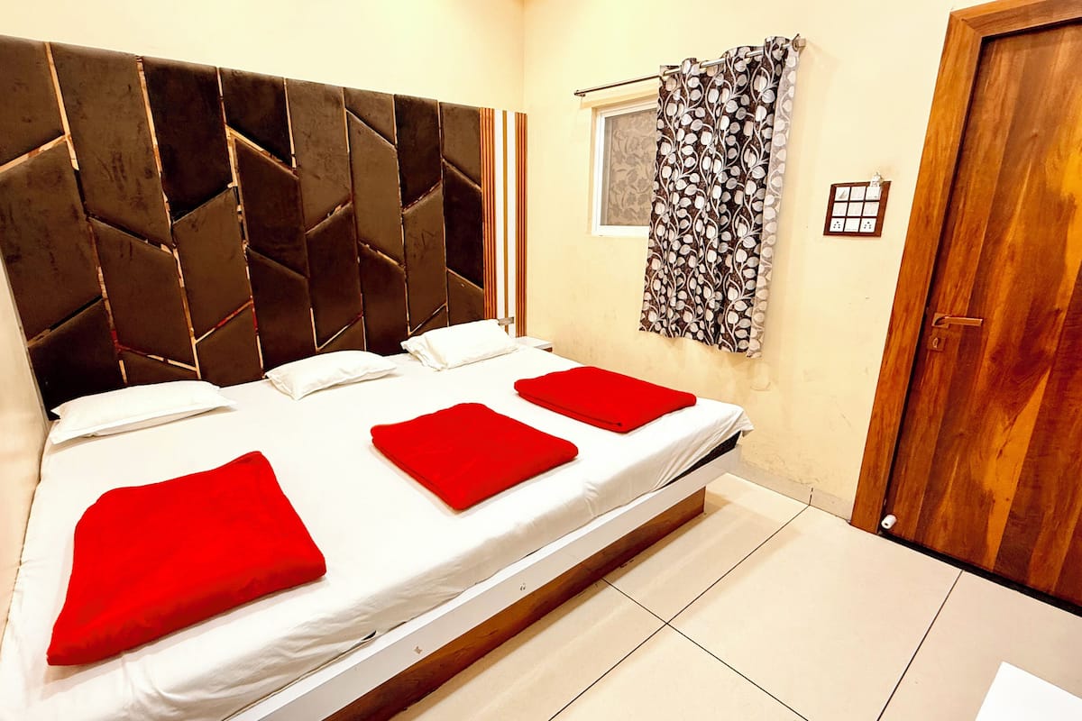 Een slaapkamer met een modern hoofdeinde, een bed met witte beddengoed en rode kussens, een raam met patroon gordijnen, en een houten deur.