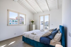 Ein Schlafzimmer mit einem Bett, einem Schminktisch, einer Vase mit Blumen und großen Fenstern.