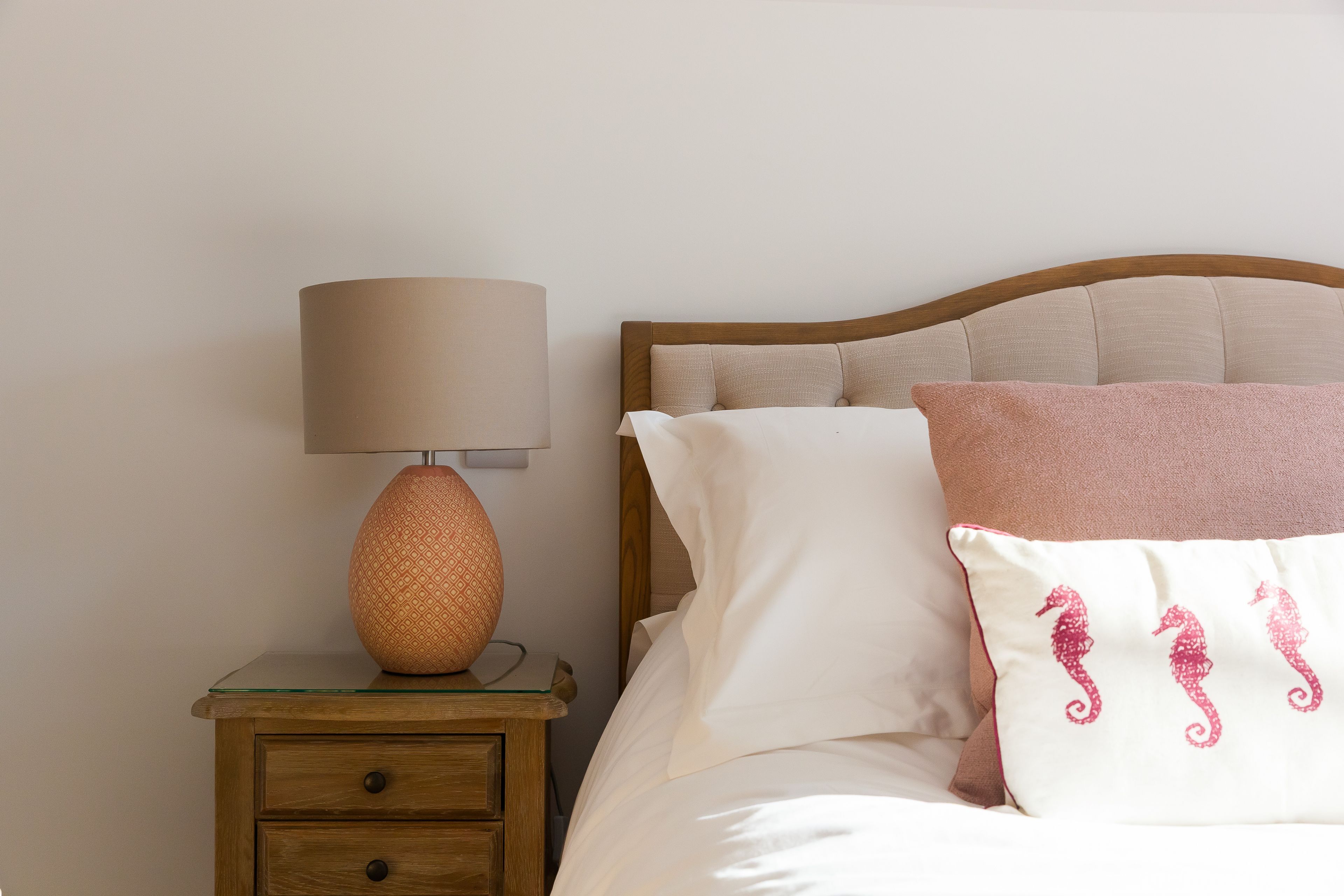 A neatly made bed with white and pink pillows, a wooden headboard, and a small bedside table with a lamp and a patterned vase.