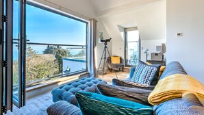 A modern living room with a large window, a cozy sofa set, and a tripod-mounted camera. - Sea Vista, Carbis Bay – Sleeps 6 – Sea Views – Penthouse – Dog-Friendly - On-Site Parking (Carbis Bay)