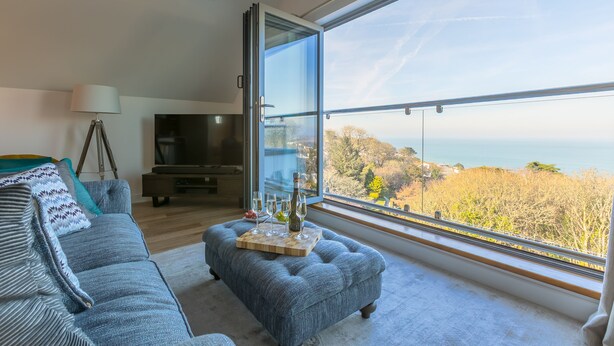 A modern living room with a large window, a grey sofa, a wooden coffee table with glasses and a bottle, and a television. - Sea Vista, Carbis Bay – Sleeps 6 – Sea Views – Penthouse – Dog-Friendly - On-Site Parking (Carbis Bay)