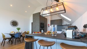A modern kitchen with a central island, bar stools, and a dining area with chairs and a table set for breakfast. - Sea Vista, Carbis Bay – Sleeps 6 – Sea Views – Penthouse – Dog-Friendly - On-Site Parking (Carbis Bay)