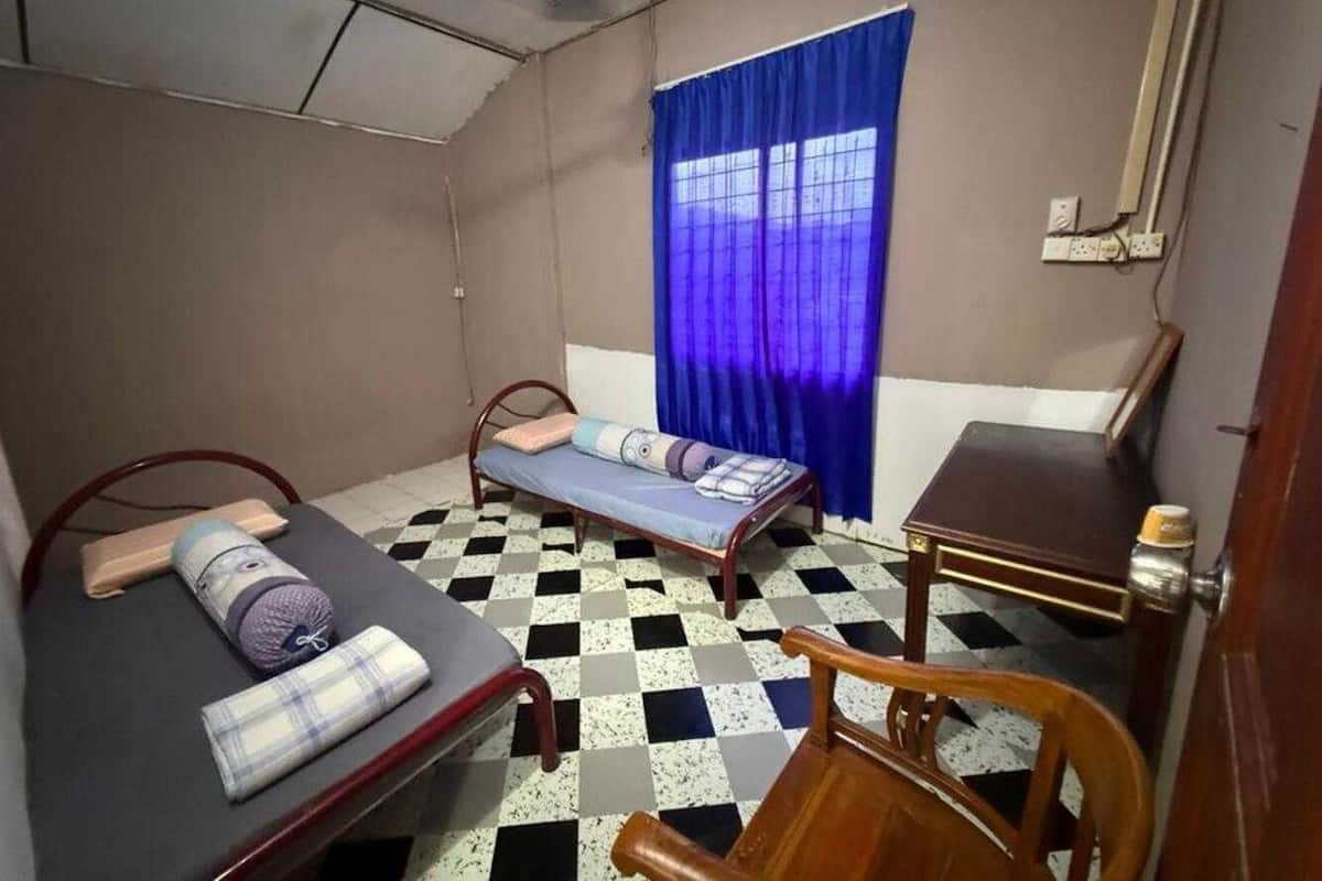 A small, compact room with two single beds, a wooden chair, a small table, and a window with blue curtains.