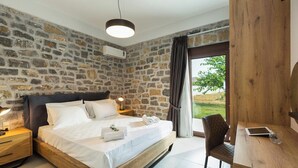 Un dormitorio con pared de piedra, una cama con ropa blanca, una mesita de noche con lámpara y vistas al verdor exterior.