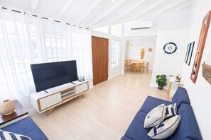 A modern living room with a flat-screen TV, a sofa, a dining area, and a large window with curtains. - Complete house in Búzios with pool and gourmet area for up to 10 people. (Armação dos Búzios)