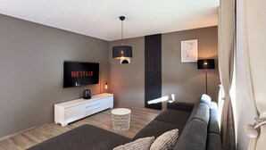A modern living room with a grey sofa, a white entertainment unit, a flat-screen TV displaying 'NETFLIX', and a pendant light. - Spacious 118m² Suite for Work & Play | 3BR, 2BA, Office, WiFi, Parking (Remchingen)