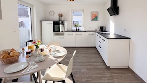 A modern kitchen with white cabinets, a black countertop, and a built-in oven. There is a dining table set with breakfast items and a vase of flowers. - Spacious 118m² Suite for Work & Play | 3BR, 2BA, Office, WiFi, Parking (Remchingen)