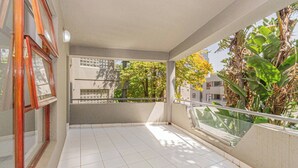 Uma varanda com piso de azulejos, guarda-corpo de vidro e vista para árvores e edifícios.
