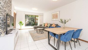 Uma sala de estar moderna com uma mesa de jantar de madeira, cadeiras azuis, um sofá cinza com almofadas coloridas, uma pequena mesa de centro, uma televisão e uma grande janela com vista para árvores.