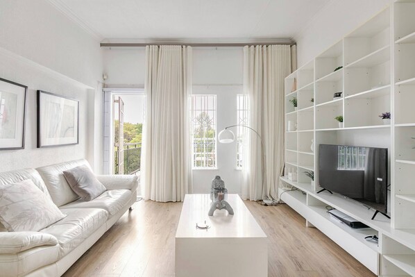 Un salon moderne avec un canapé blanc, une table basse, un téléviseur posé sur un meuble blanc et un balcon visible à travers les fenêtres.
