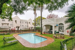 A swimming pool with a brick border, surrounded by a grassy area with lounge chairs. There are palm trees and a building with a white facade in the background. - Cosy 2bed in Heart of Sandton (Johannesburg)