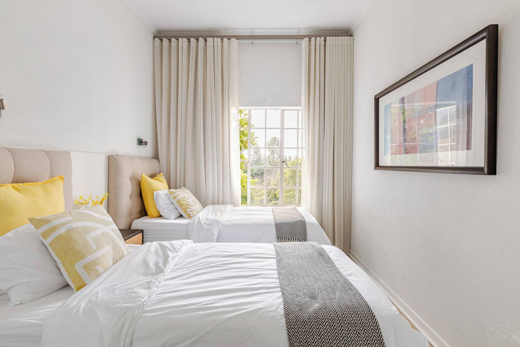 Une chambre d’hôtel avec deux lits, une grande fenêtre avec des rideaux, un tableau encadré au mur et une vue sur la verdure.