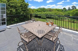 Un balcon avec une table et des chaises en métal, un pot de fleurs et une vue sur un paysage verdoyant.