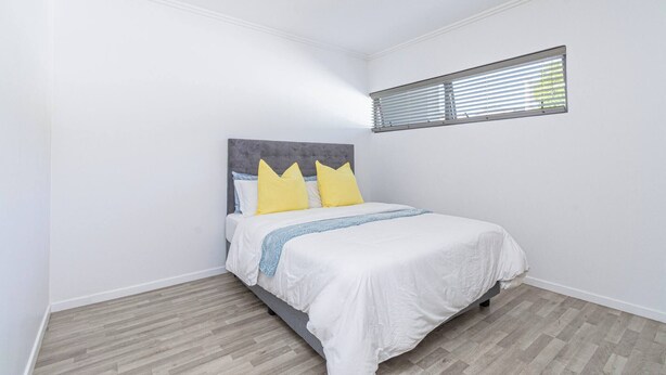 Un dormitorio con una cama, paredes blancas, un ventanal con persianas y suelo de madera.