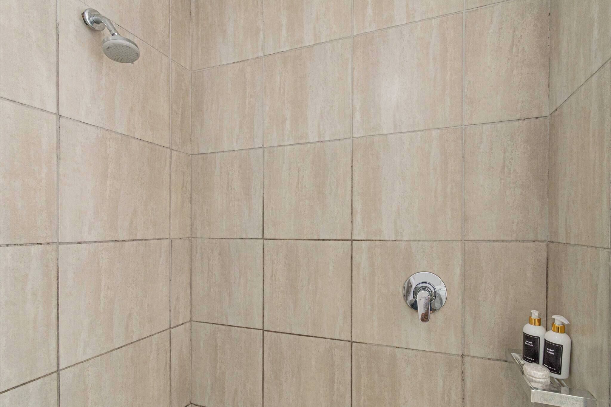 A shower area with beige tiles, a handheld showerhead, a wall-mounted faucet, and two bottles of shampoo on a shelf.