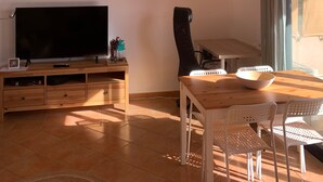 A modern living room with a flat-screen TV, wooden entertainment unit, white chairs, a woven pendant light, and a dining table.