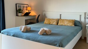A neatly made bed with towels, a desk with a lamp, and a hanging light fixture in a hotel room.