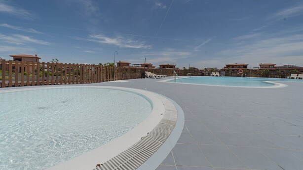 A swimming pool area with multiple pools, a wooden fence, and buildings in the background.