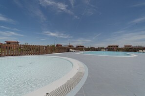A swimming pool area with multiple pools, a wooden fence, and buildings in the background.