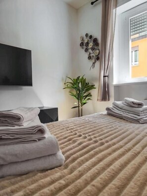 Una cama bien hecha con una manta beige con textura, una pila de toallas dobladas, una planta en maceta y un televisor fijado a la pared.