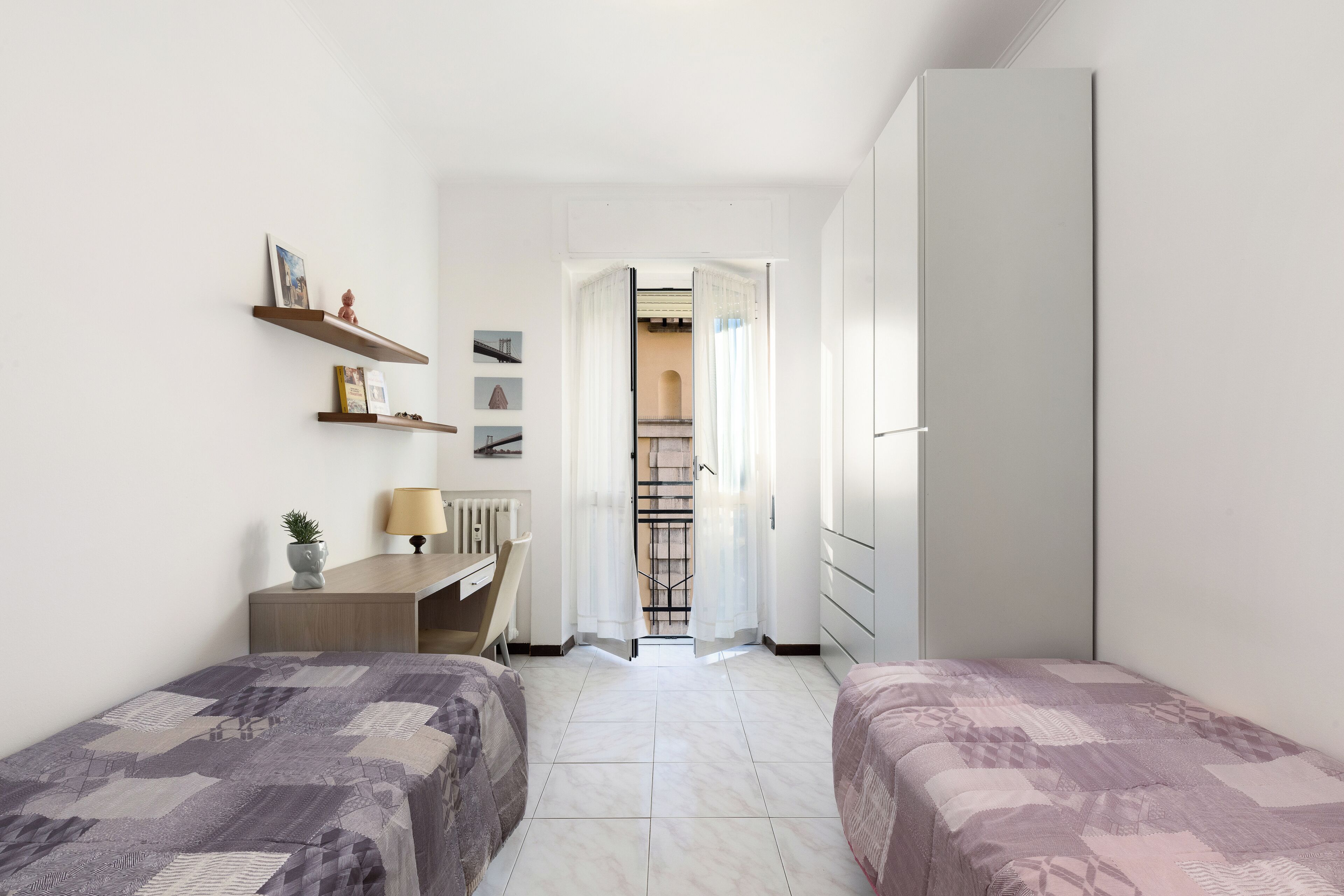 Une petite chambre bien éclairée, avec deux lits, un bureau et une armoire.