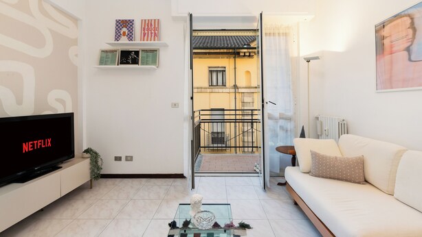 A modern living room with a flat-screen TV, a sofa, a coffee table with decorative items, and a view of a balcony through an open door. - Large Apartment Sesto Rondò M1 - Duomo 20 min (Sesto San Giovanni)