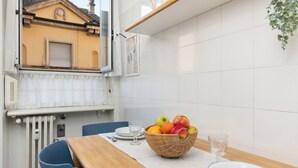 A dining area with a table set for two, a wicker fruit bowl, and a window overlooking a building. - Large Apartment Sesto Rondò M1 - Duomo 20 min (Sesto San Giovanni)