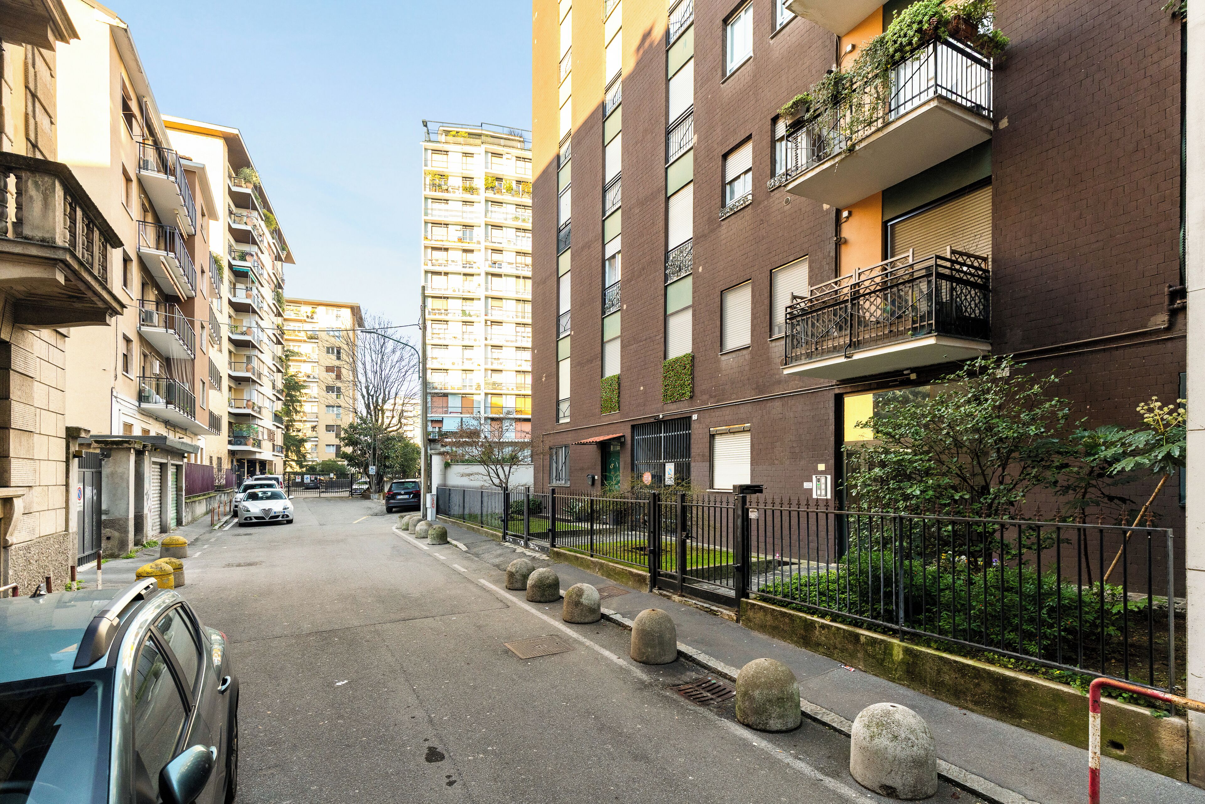 Une rue étroite en milieu urbain, bordée d’immeubles résidentiels à plusieurs étages, avec des balcons ornés de plantes, un ciel dégagé et une voiture garée.