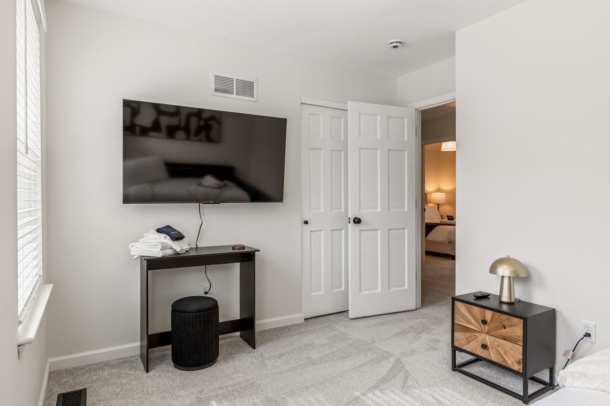 A modern bedroom with a TV mounted on the wall, a bedside table with a lamp, and a door leading to another room.