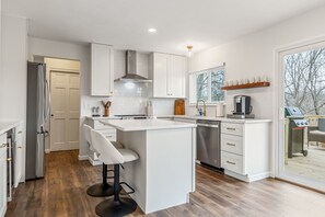 A modern kitchen with white cabinetry, a central island with bar stools, a dishwasher, and a view of the outdoors through a sliding glass door.