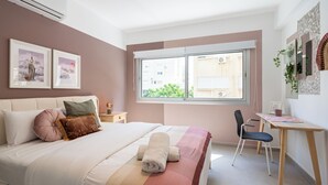 A bedroom with a bed, a desk, and a chair. There is a window with a view of buildings outside. - Cloudwalk 1- BR Apartment in Larnaca (Larnaca)