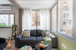 Ein modernes Wohnzimmer mit einem grauen Sofa, einem kleinen Couchtisch und Blick auf ein Gebäude durch ein großes Fenster.
