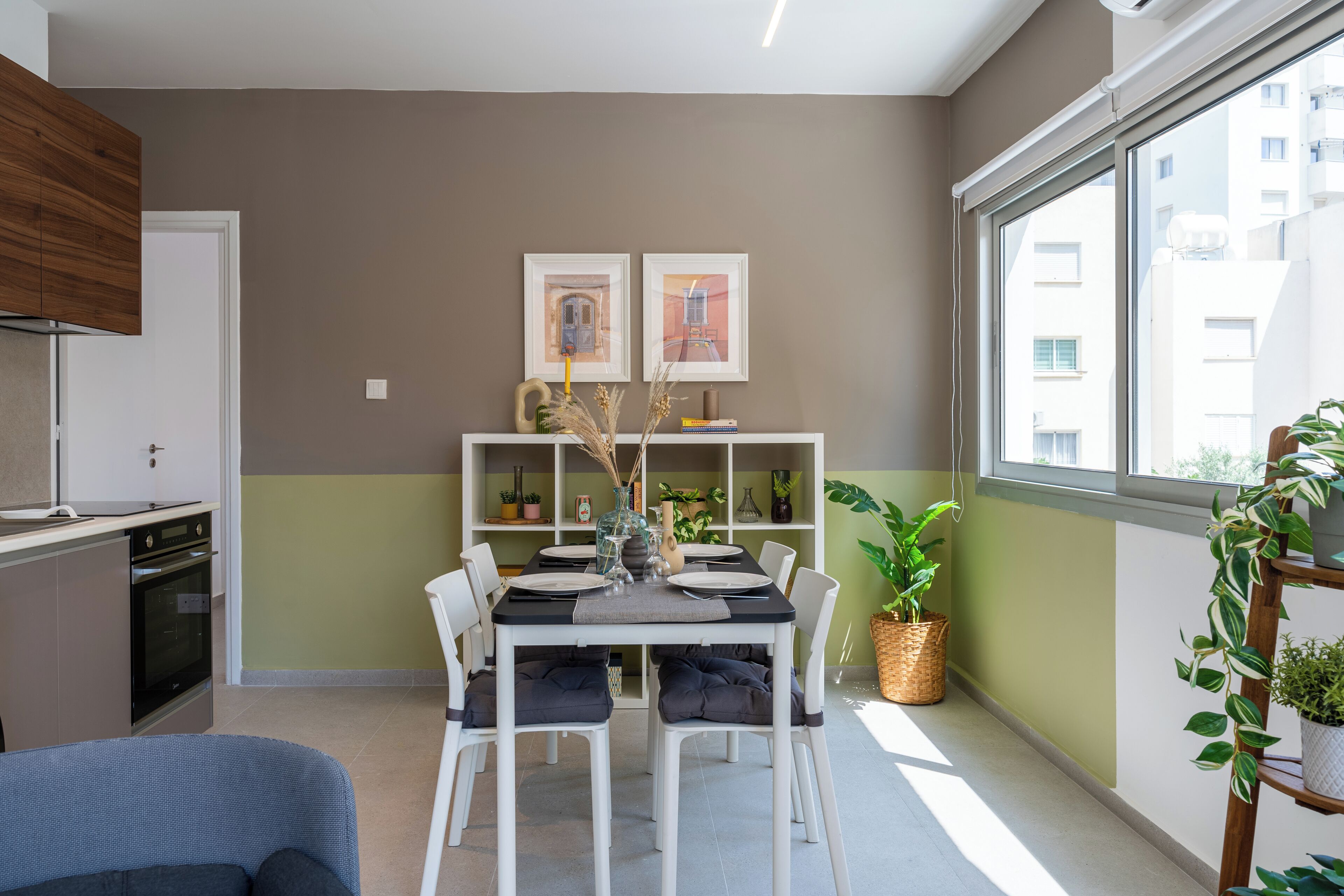 Une cuisine moderne avec des armoires blanches, un four encastré et un coin repas avec une table dressée pour quatre personnes. Deux tableaux encadrés ornent le mur, ainsi qu’une plante en pot et une fenêtre donnant sur des immeubles.
