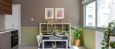 Une cuisine moderne avec des armoires blanches, un four encastré et un coin repas avec une table dressée pour quatre personnes. Deux tableaux encadrés ornent le mur, ainsi qu’une plante en pot et une fenêtre donnant sur des immeubles.