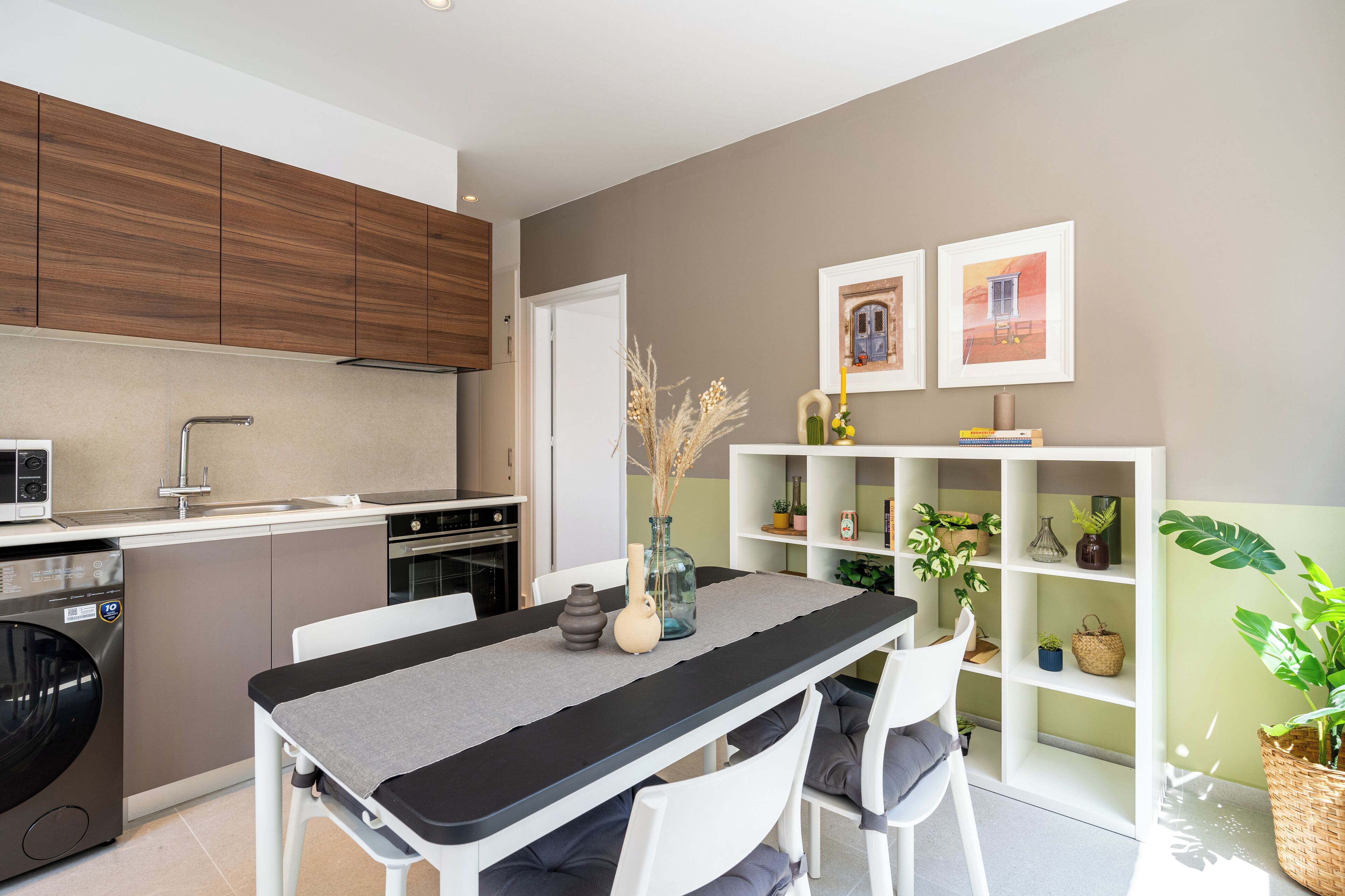A modern kitchen with a dining area, white cabinetry, and a built-in bookshelf with plants.