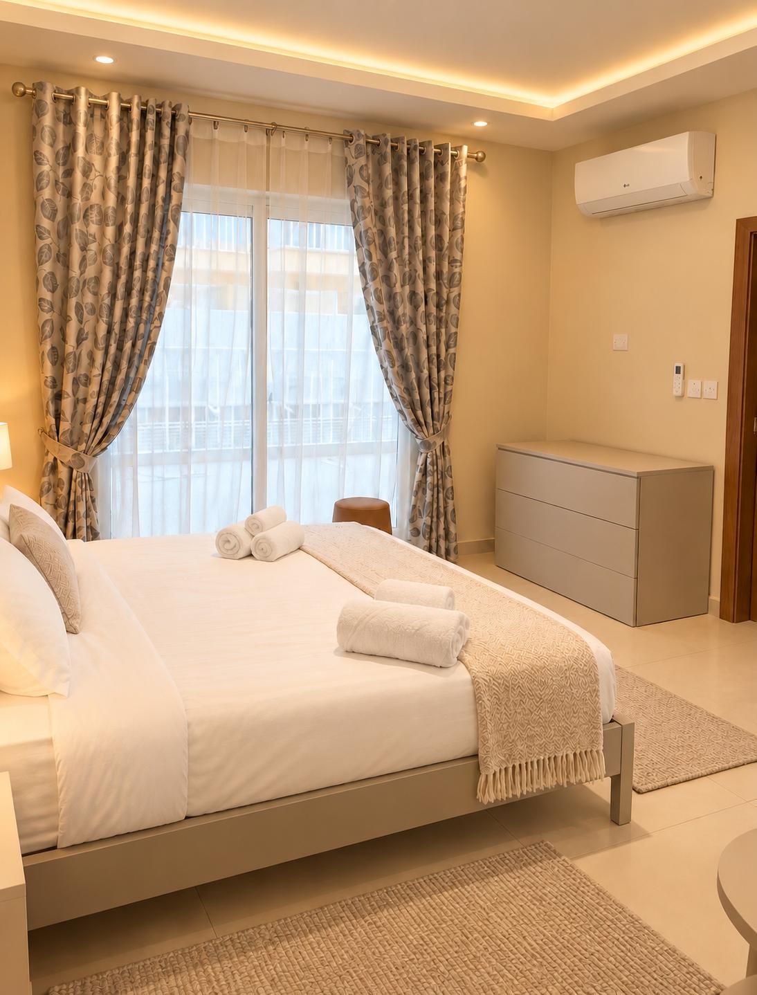 A modern bedroom with a bed, curtains, a bedside table, a dresser, and an air conditioning unit.
